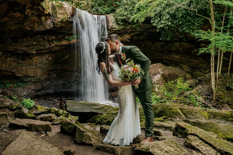 James and Emily kissing at a waterfall on their wedding day, surrounded by wedding flowers