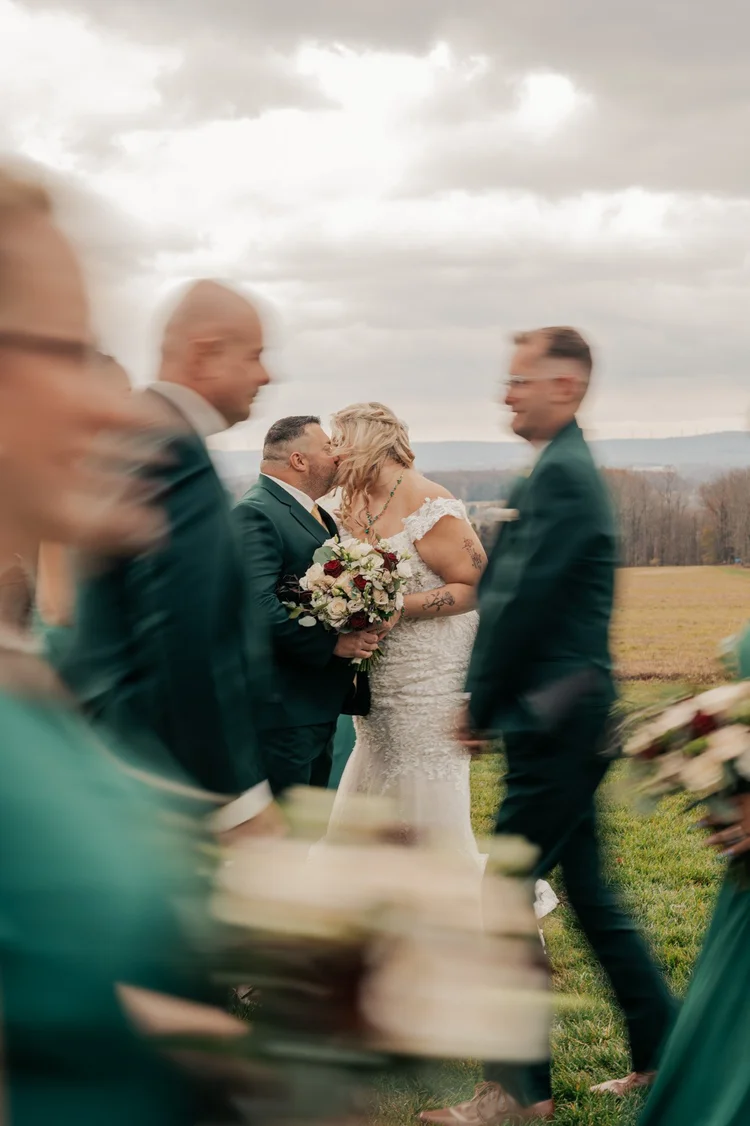 A bride and groom share a kiss outdoors during their wedding, surrounded by wedding guests