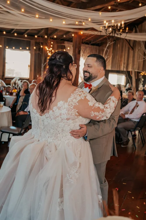 A bride and groom dancing at their wedding reception inside a rustic barn with string lights